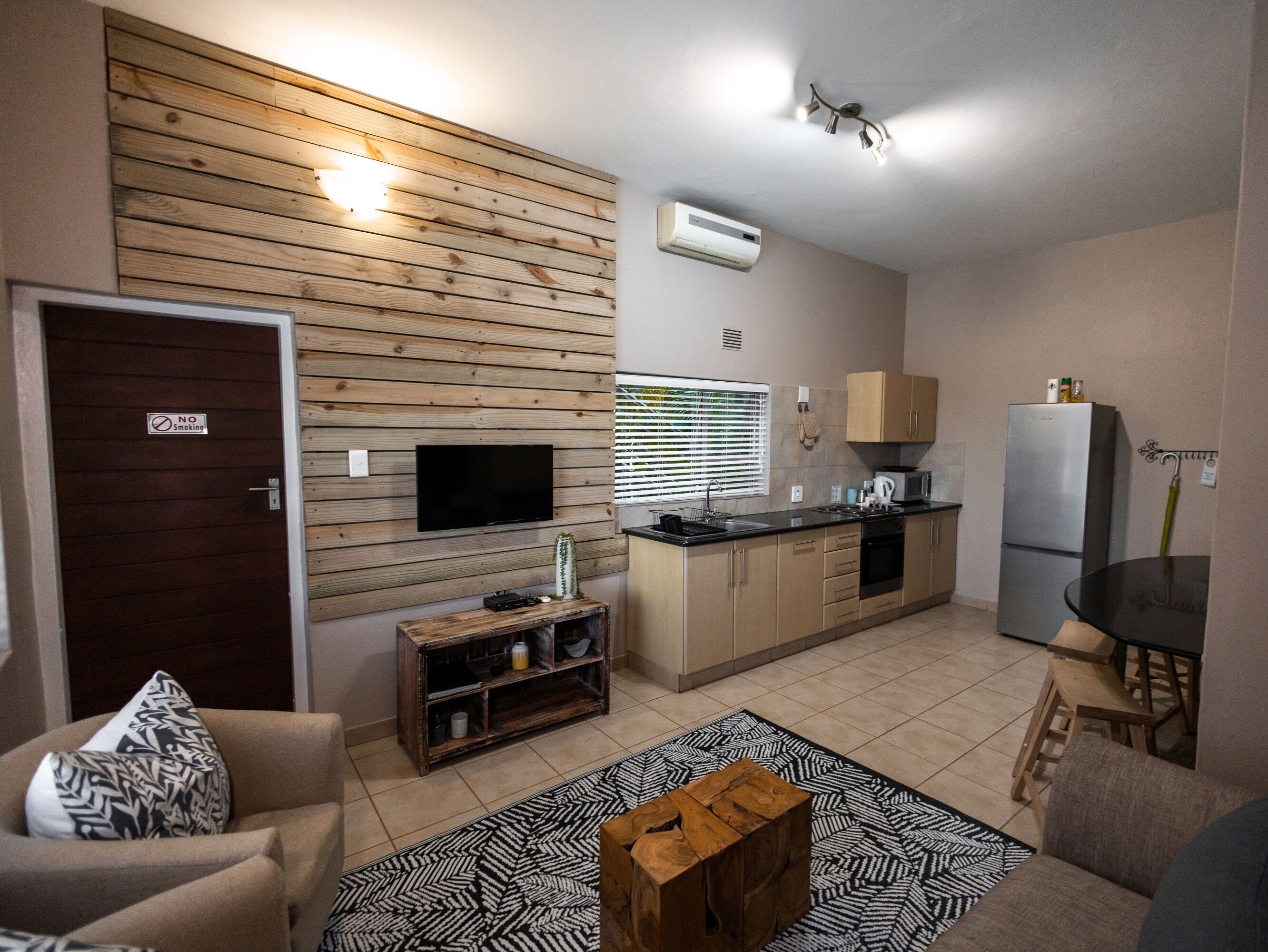 A compact living space with a kitchenette, a flat-screen TV mounted on a wooden wall, a dining area with a table and chairs, and a seating area with a sofa and a patterned rug.