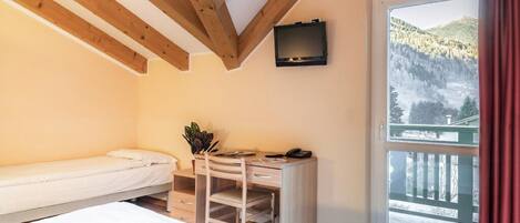 A bedroom with a bed, a desk with a chair, a TV, and a balcony with a view of a forested mountain.