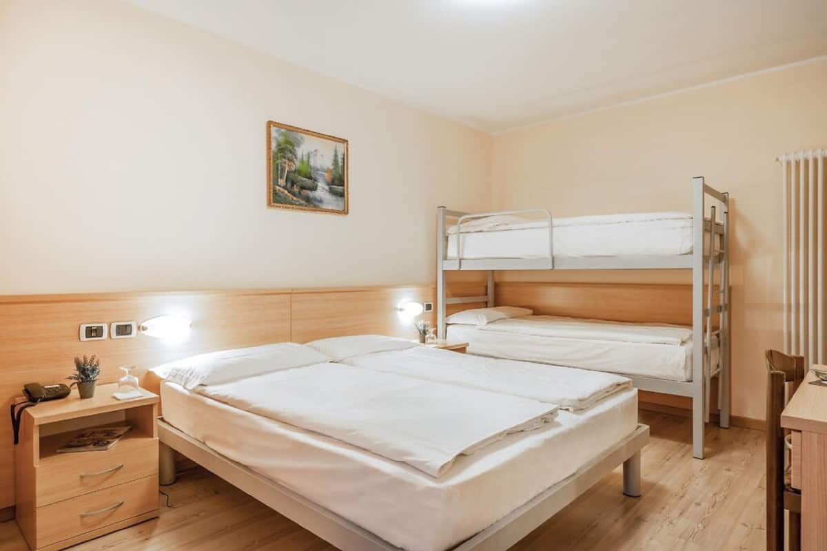 A hotel room with a single bed, a bunk bed, wooden furniture, a bedside table with a lamp, and a painting on the wall.