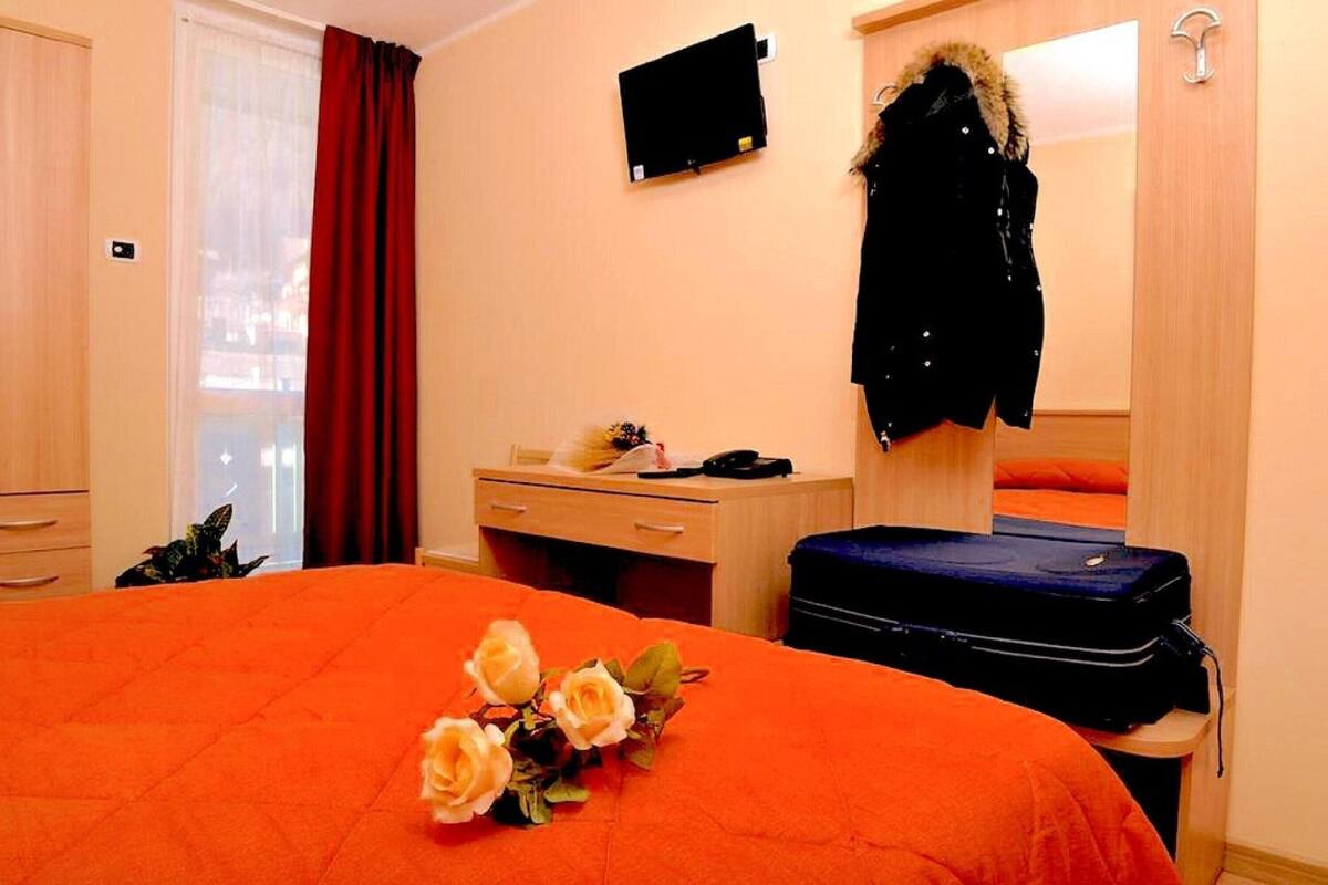 A hotel room with a bed, a desk, a chair, a suitcase, and a coat hanging on the wall.
