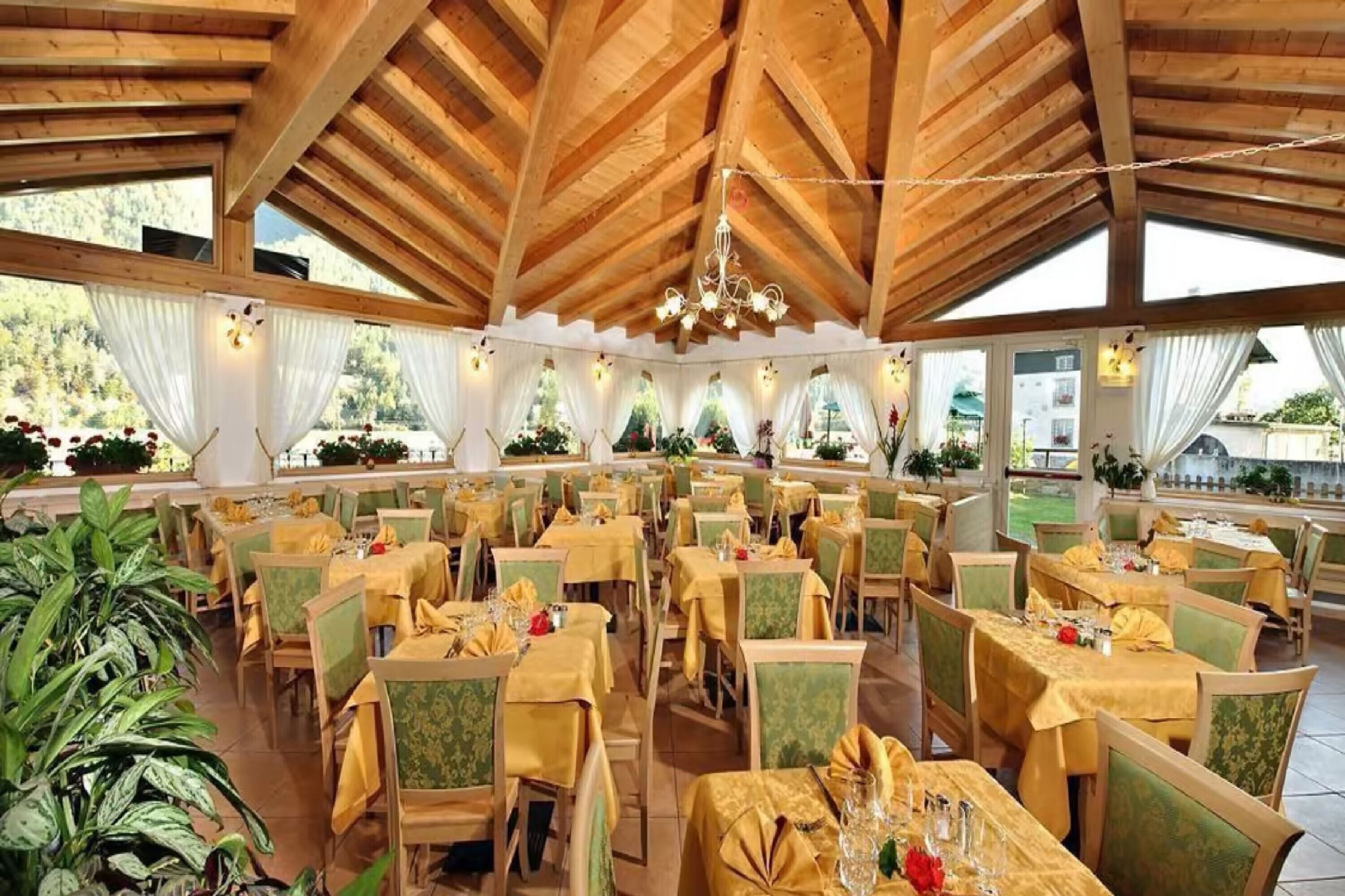 Un'ampia sala da pranzo con travi in legno e ampie finestre, tavoli apparecchiati con tovaglie gialle e sedie verdi.