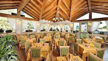 Un'ampia sala da pranzo con travi in legno e ampie finestre, tavoli apparecchiati con tovaglie gialle e sedie verdi.