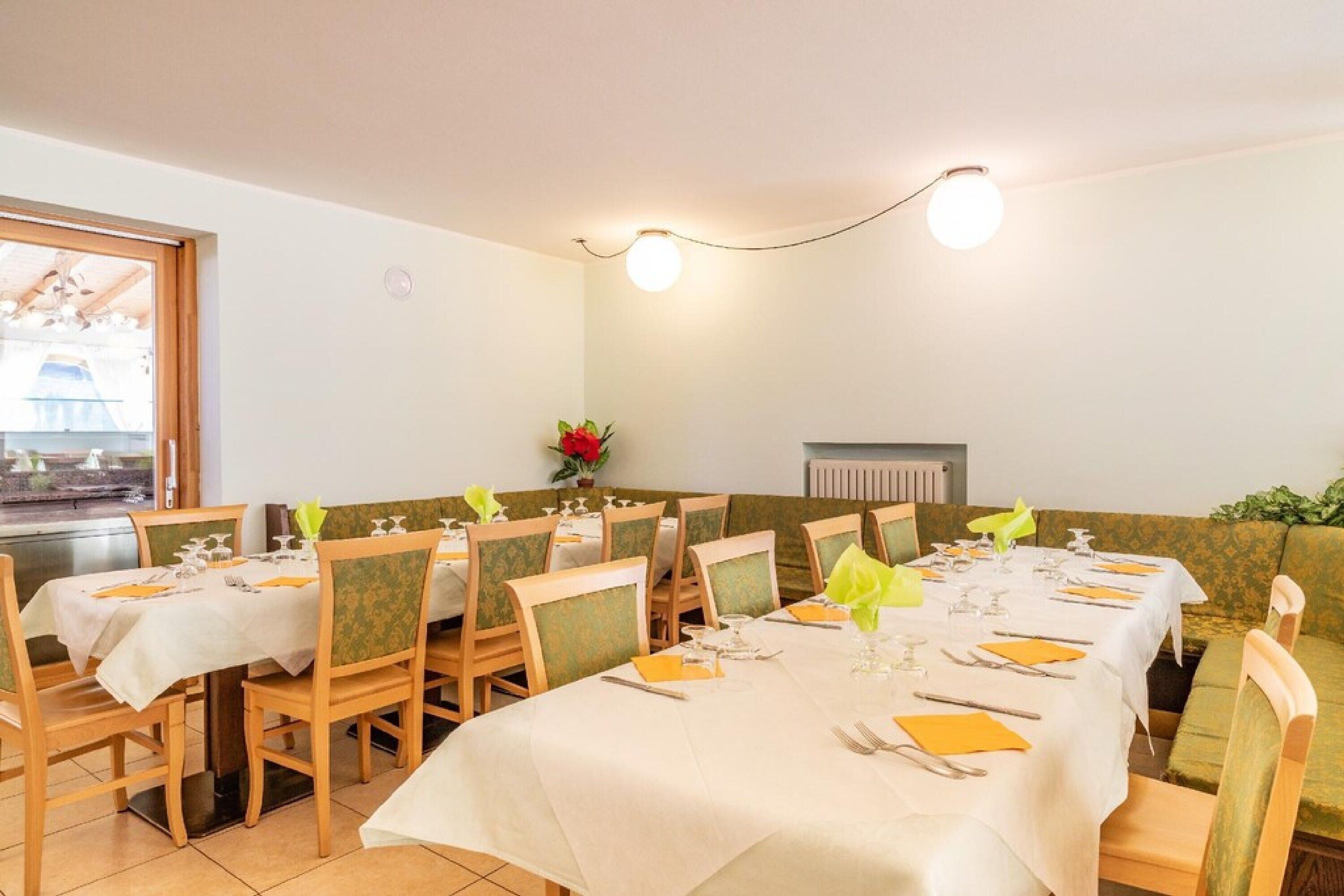 A dining area with tables set for four, each with a white tablecloth, yellow napkins, and silverware. There are green upholstered chairs with a floral pattern. A flower vase with red flowers is on the table, and a radiator is visible on the wall.