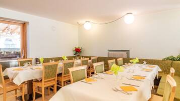 Comedor con mesas dispuestas para cuatro, cada una con mantel blanco, servilletas amarillas y cubertería. Sillas tapizadas en verde con estampado floral. Un florero con flores rojas está sobre la mesa, y se observa un radiador en la pared.