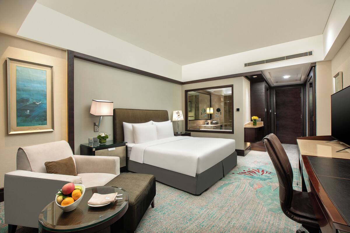 A hotel room with a bed, a desk, a chair, a sofa, a coffee table with fruit, a lamp, a painting, and a mirror.