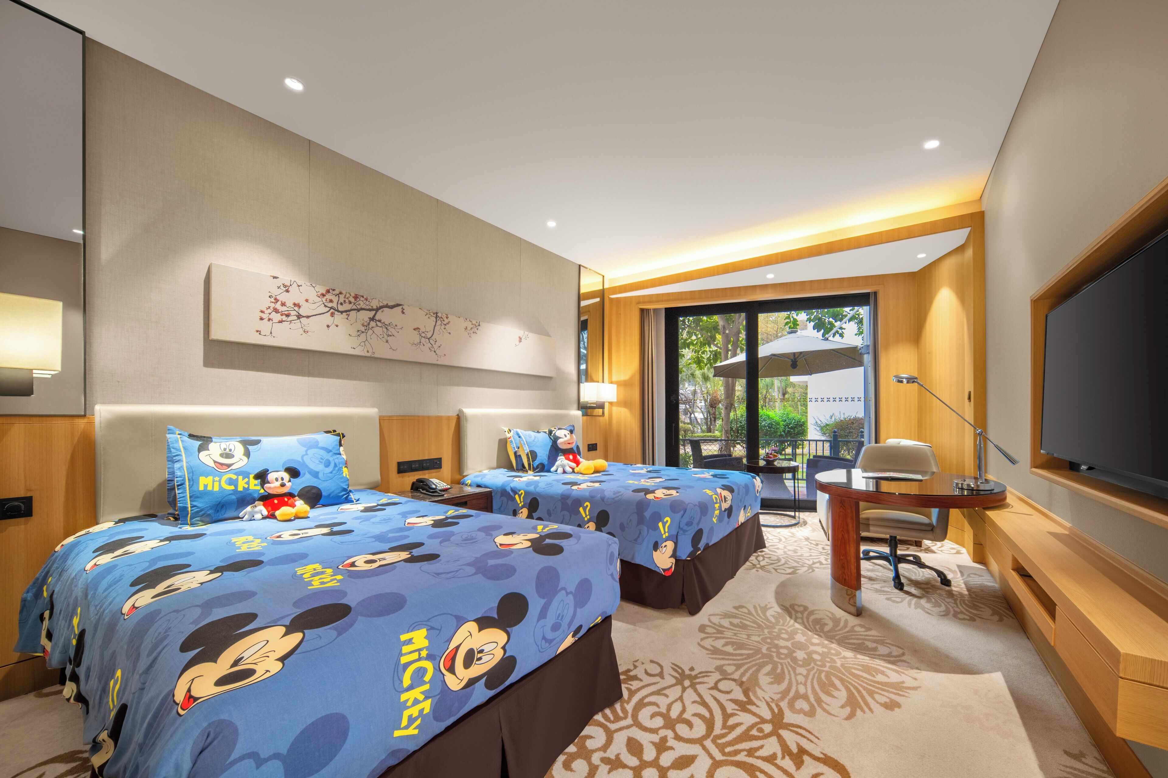 A hotel room with two beds, each with Mickey Mouse-themed bedding, a desk with a chair, a large flat-screen TV, and a window with a view of greenery.