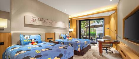 A hotel room with two beds, each with Mickey Mouse-themed bedding, a desk with a chair, a large flat-screen TV, and a window with a view of greenery.