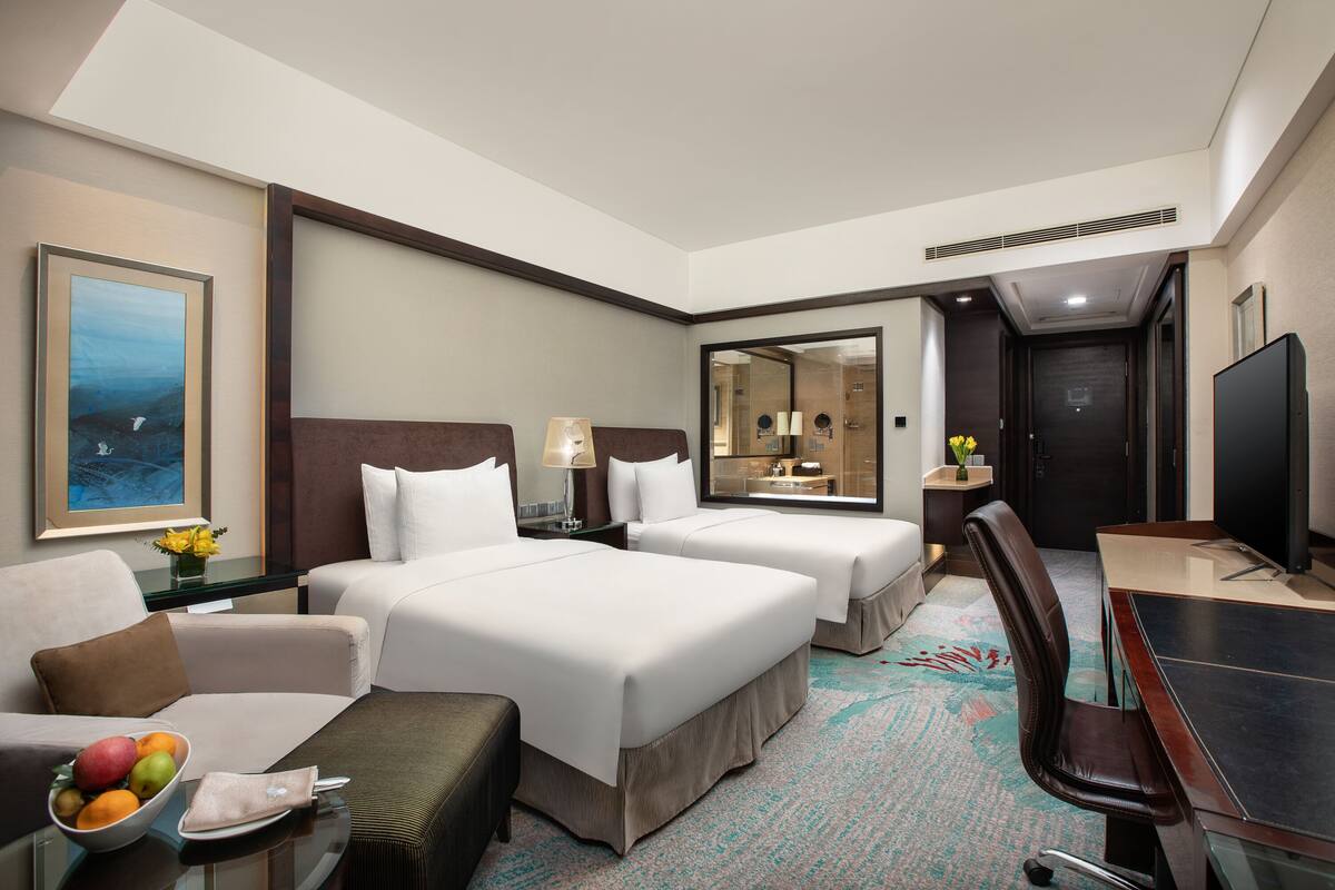 A hotel room with two beds, a desk, a chair, a TV, a mirror, and a small table with fruit.