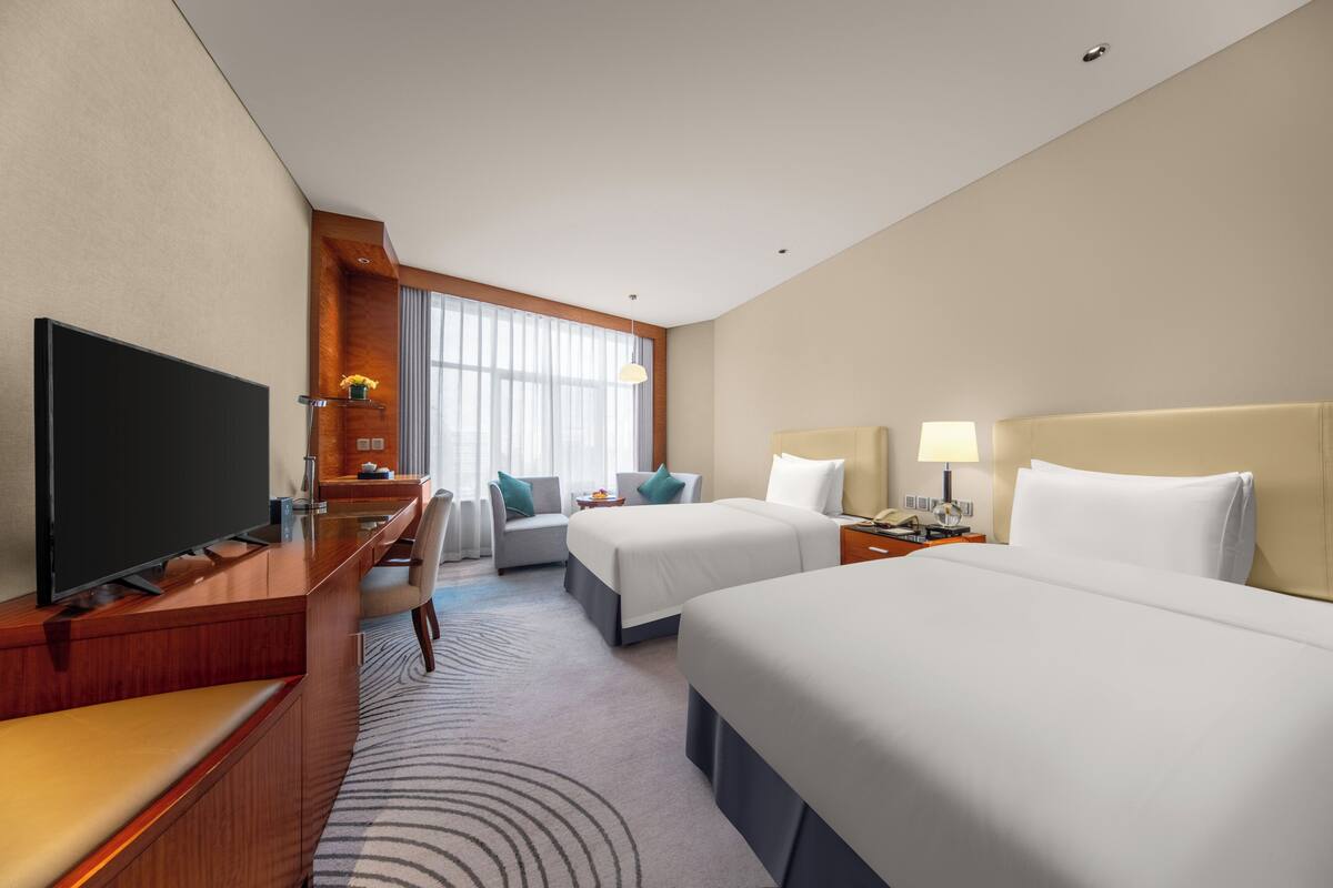 A hotel room with two beds, a desk with a TV, a chair, a lamp, and a window with curtains.