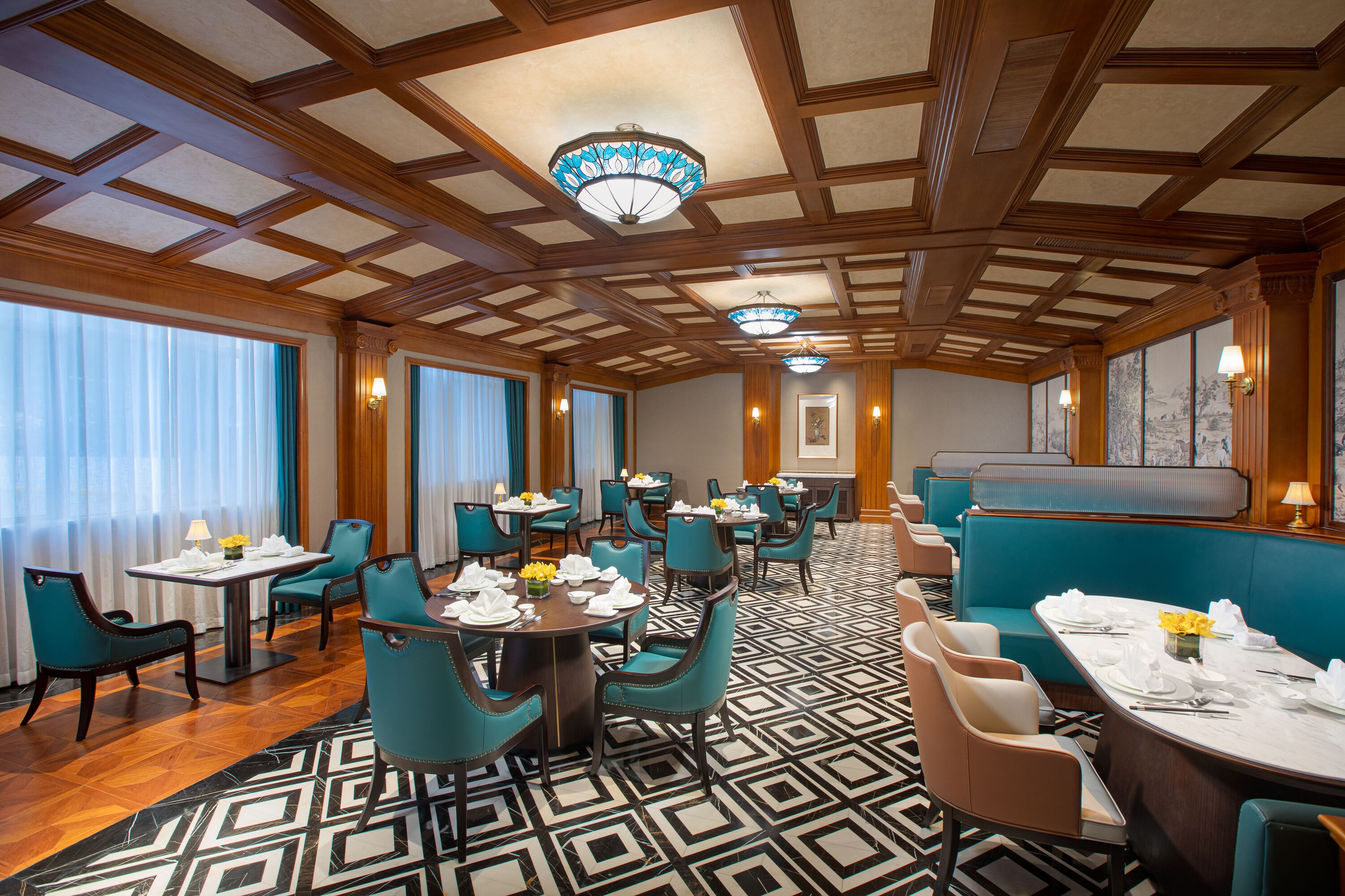 A hotel dining area with multiple tables and chairs, a patterned floor, and a ceiling with wooden beams and chandeliers.