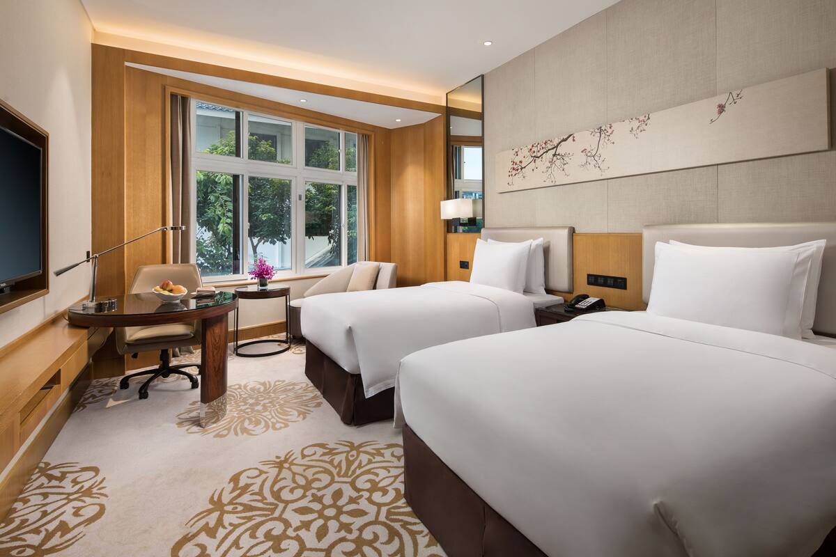 A hotel room with two beds, a desk with a chair, a TV, and a window with a view of greenery.