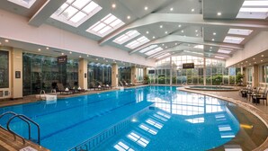 Una amplia piscina cubierta con paredes de vidrio transparente, varias zonas de descanso y un alto techo con tragaluces.