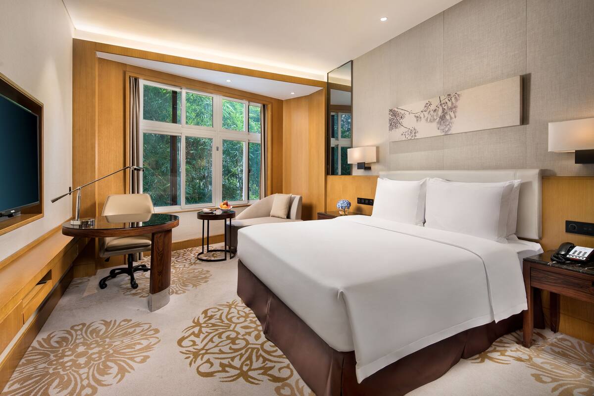 A hotel room with a large bed, a desk with a chair, a TV, and a window with a view of greenery.