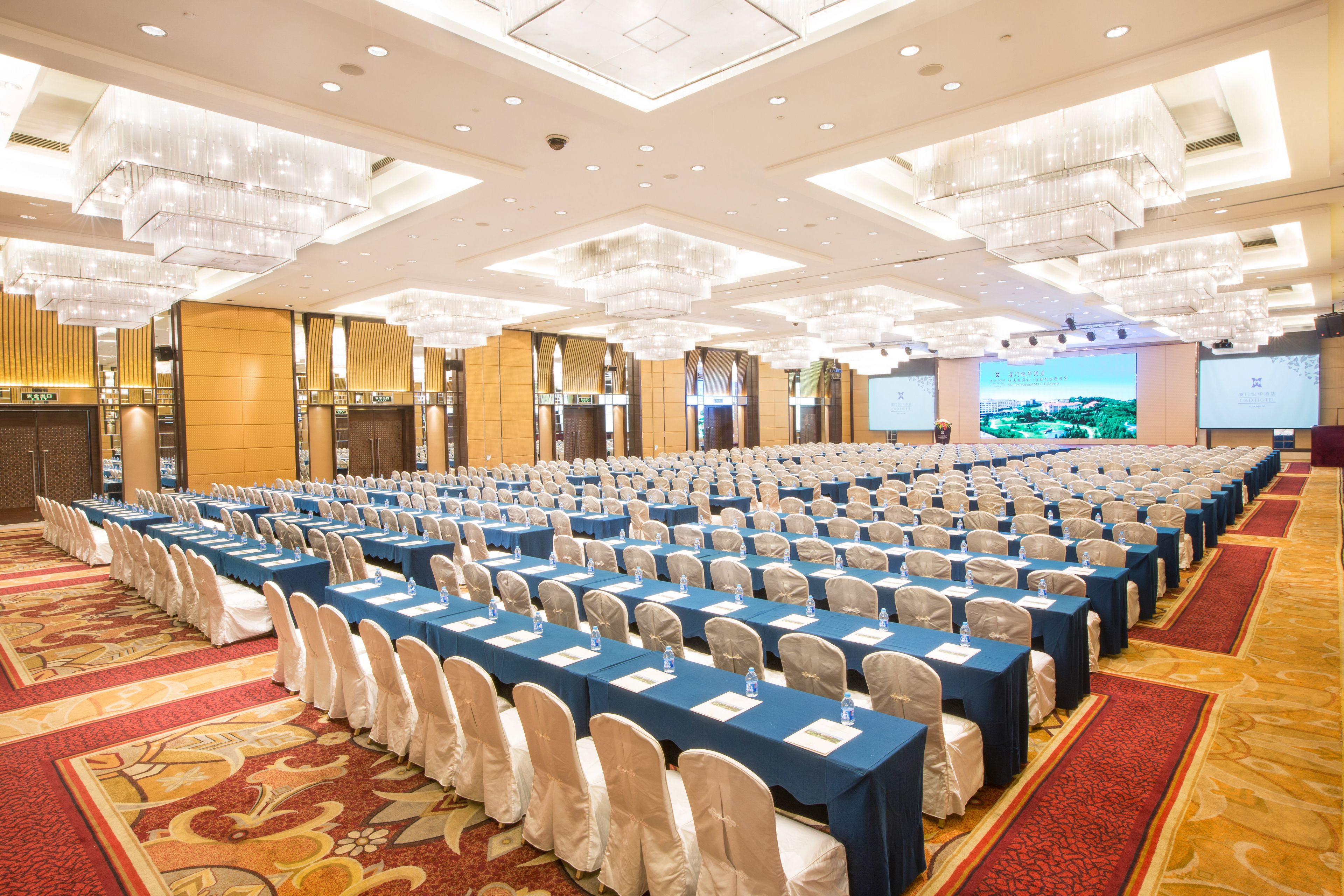 A conference room with rows of tables and chairs, a large screen, and a carpeted floor.