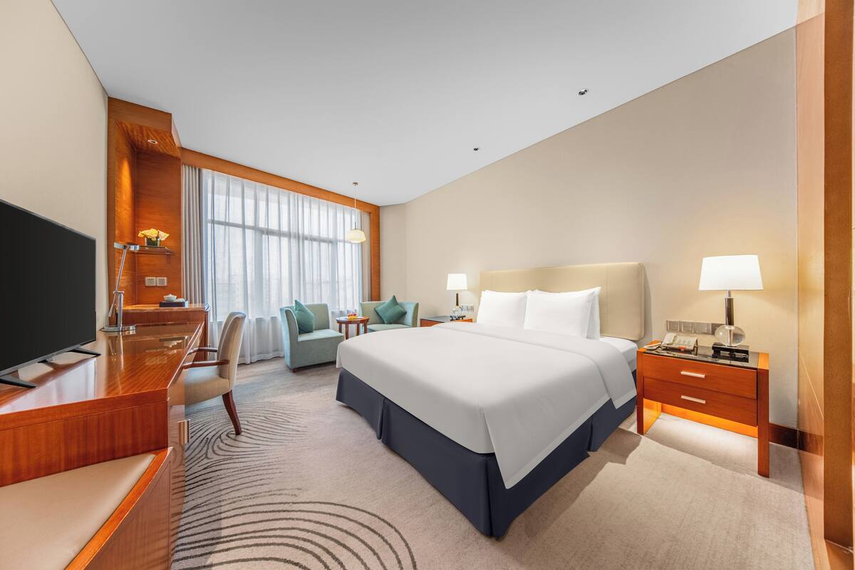 A hotel room with a large bed, a desk with a flat-screen TV, a chair, a small table, and a nightstand with a lamp.