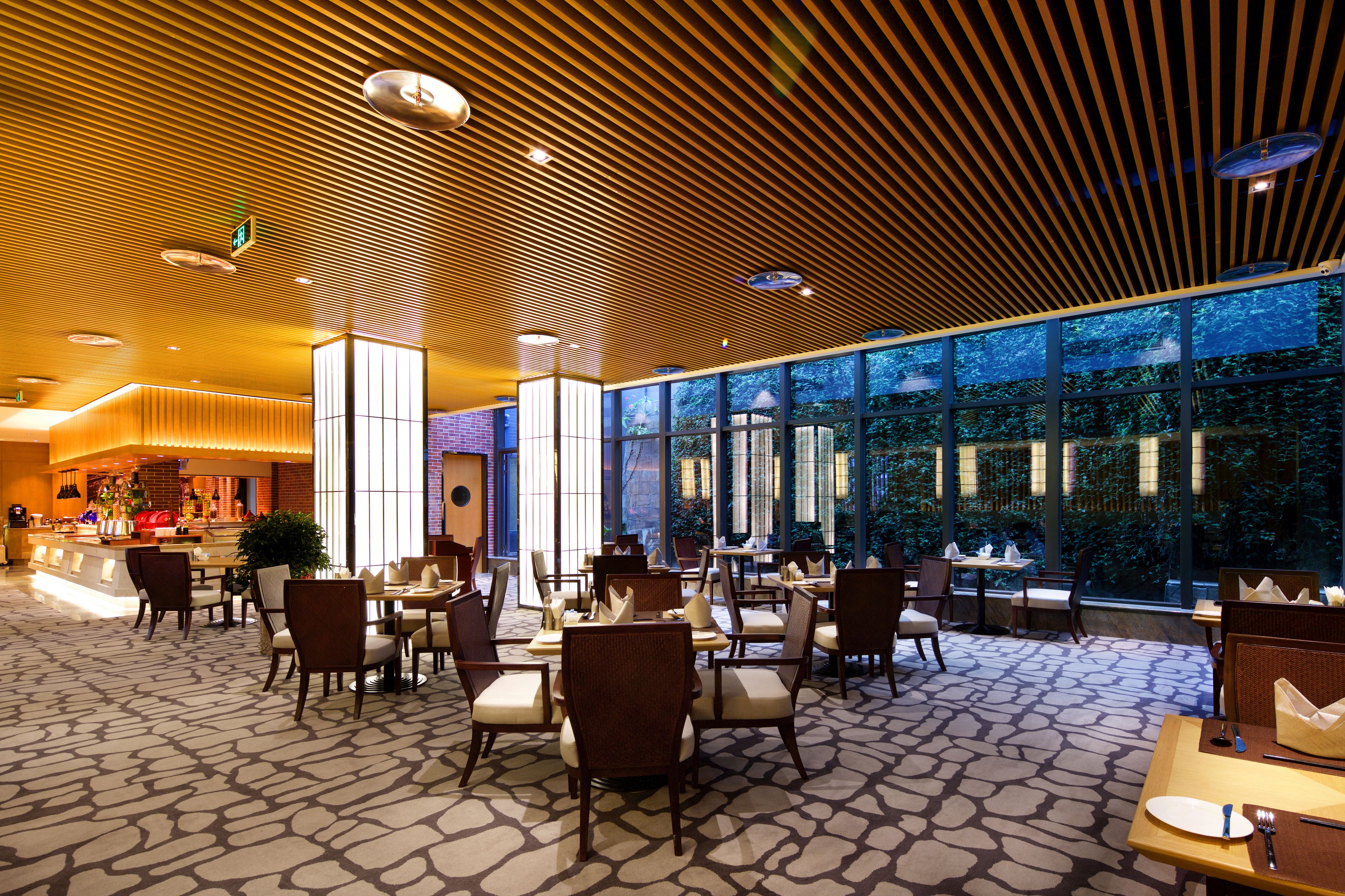 A spacious hotel lobby with a modern design, featuring a patterned carpet, wooden ceiling, and large windows offering a view of the outdoors.