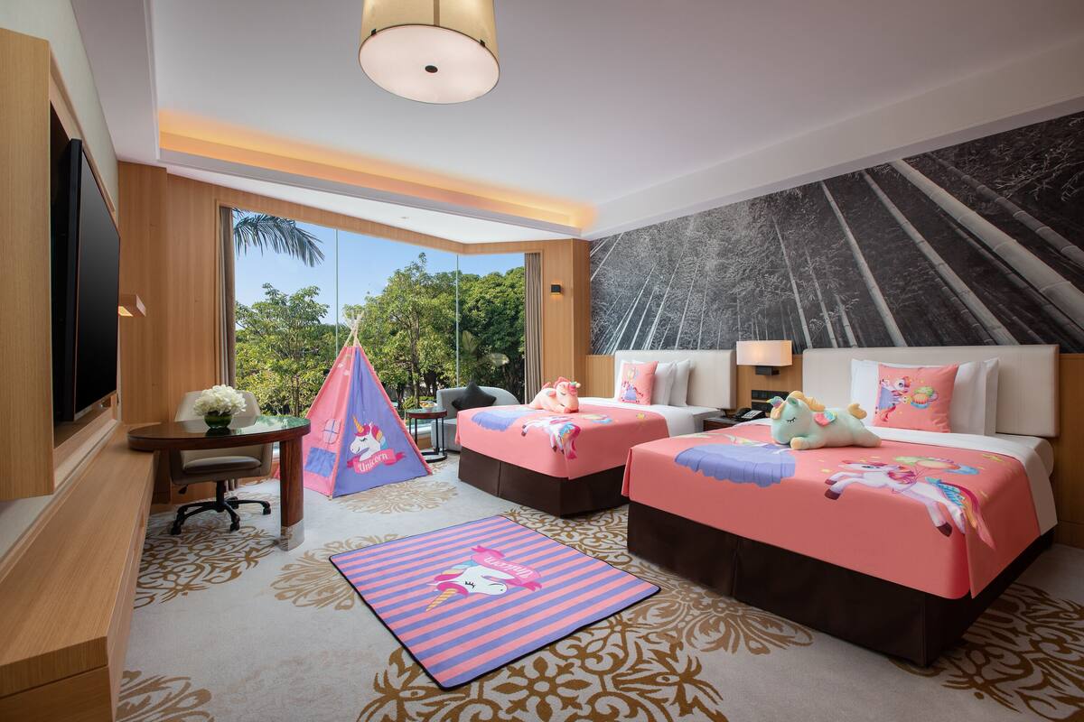 A hotel room with two beds, a desk, a chair, a small table, a hanging chair, and a large window with a view of greenery.