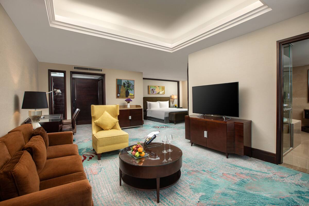 A hotel room with a brown sofa, a yellow armchair, a round coffee table with fruit and glasses, a flat-screen TV, and a large mirror.