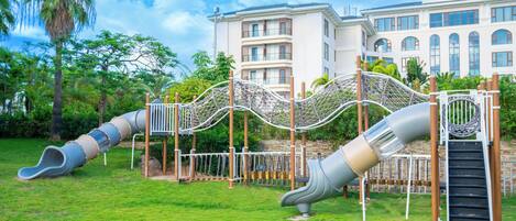 Un parque infantil con toboganes y estructuras para escalar, rodeado de un césped cuidado y un edificio grande al fondo.
