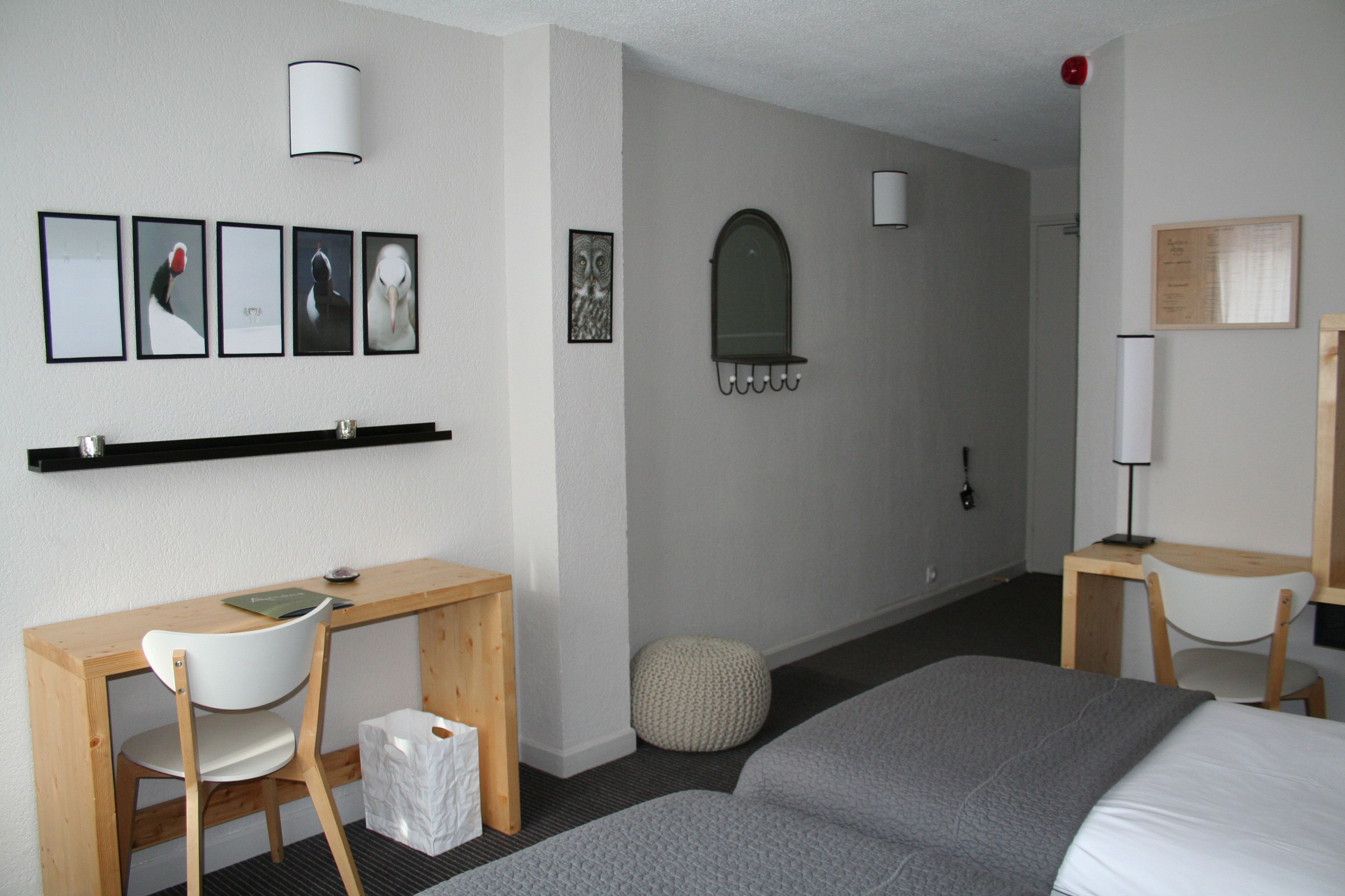 A hotel room with a bed, a desk with a chair, a small round ottoman, a wall-mounted mirror, and framed pictures on the wall.