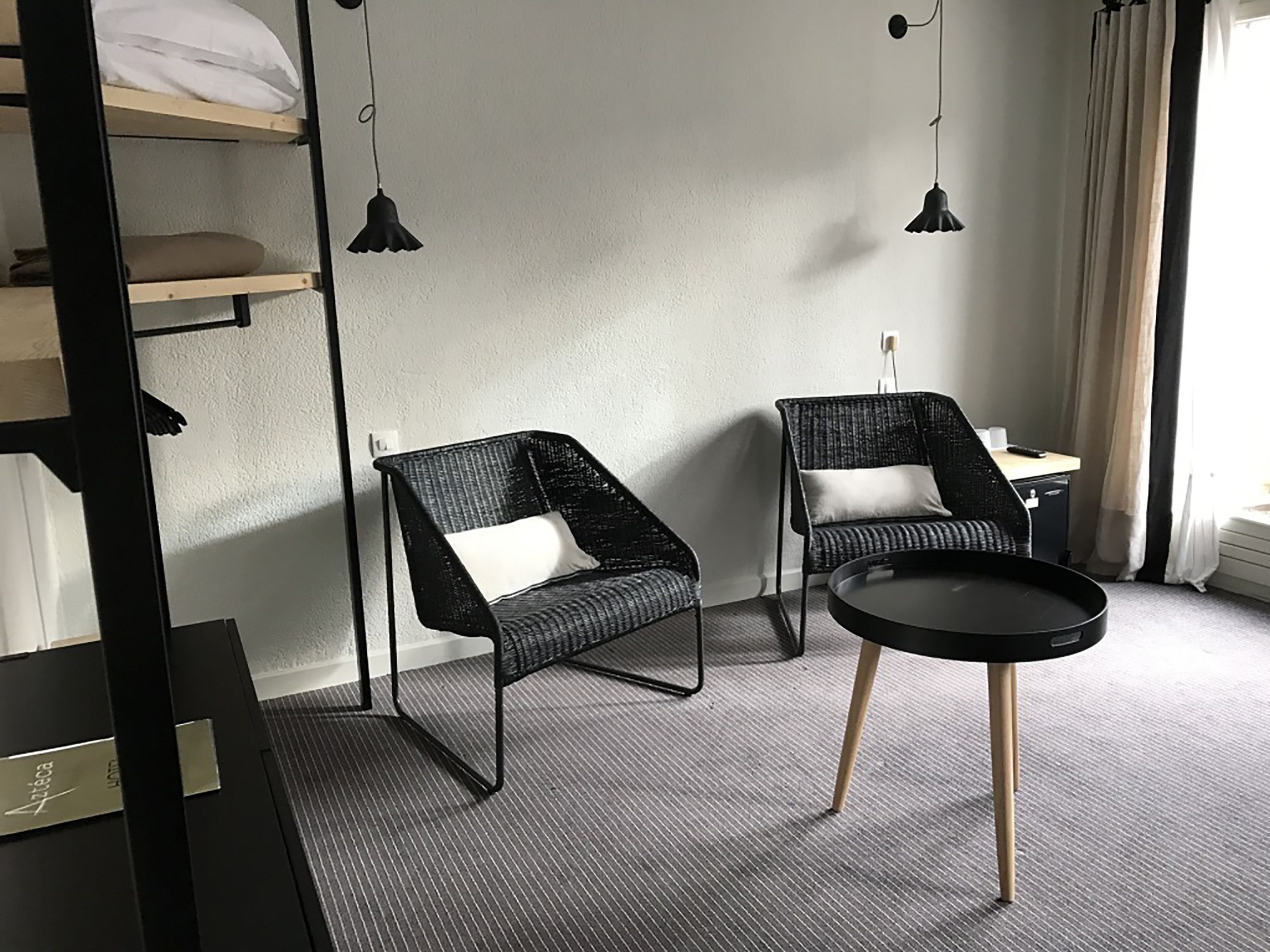 Ein modernes Hotelzimmer mit zwei schwarzen Korbstühlen, einem runden schwarzen Tisch und einem Bett mit weißer Bettwäsche.