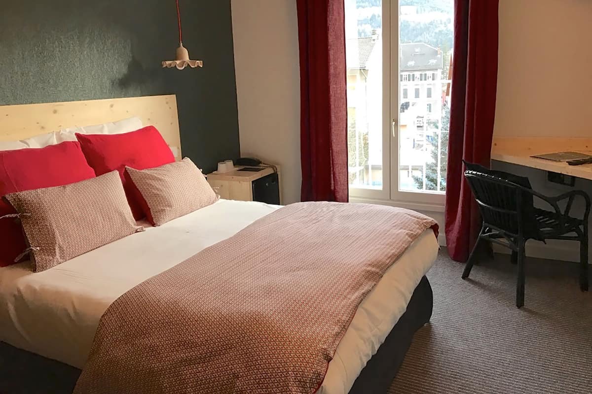 A hotel room with a bed, a desk, and a chair. There is a window with curtains, and a view of buildings outside.