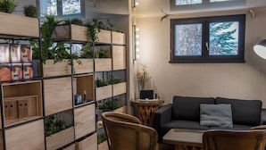 A cozy living area with a dark sofa, wicker chairs, a small wooden table, and a built-in shelving unit with plants and storage boxes. - Hotel Azteca (Barcelonnette)