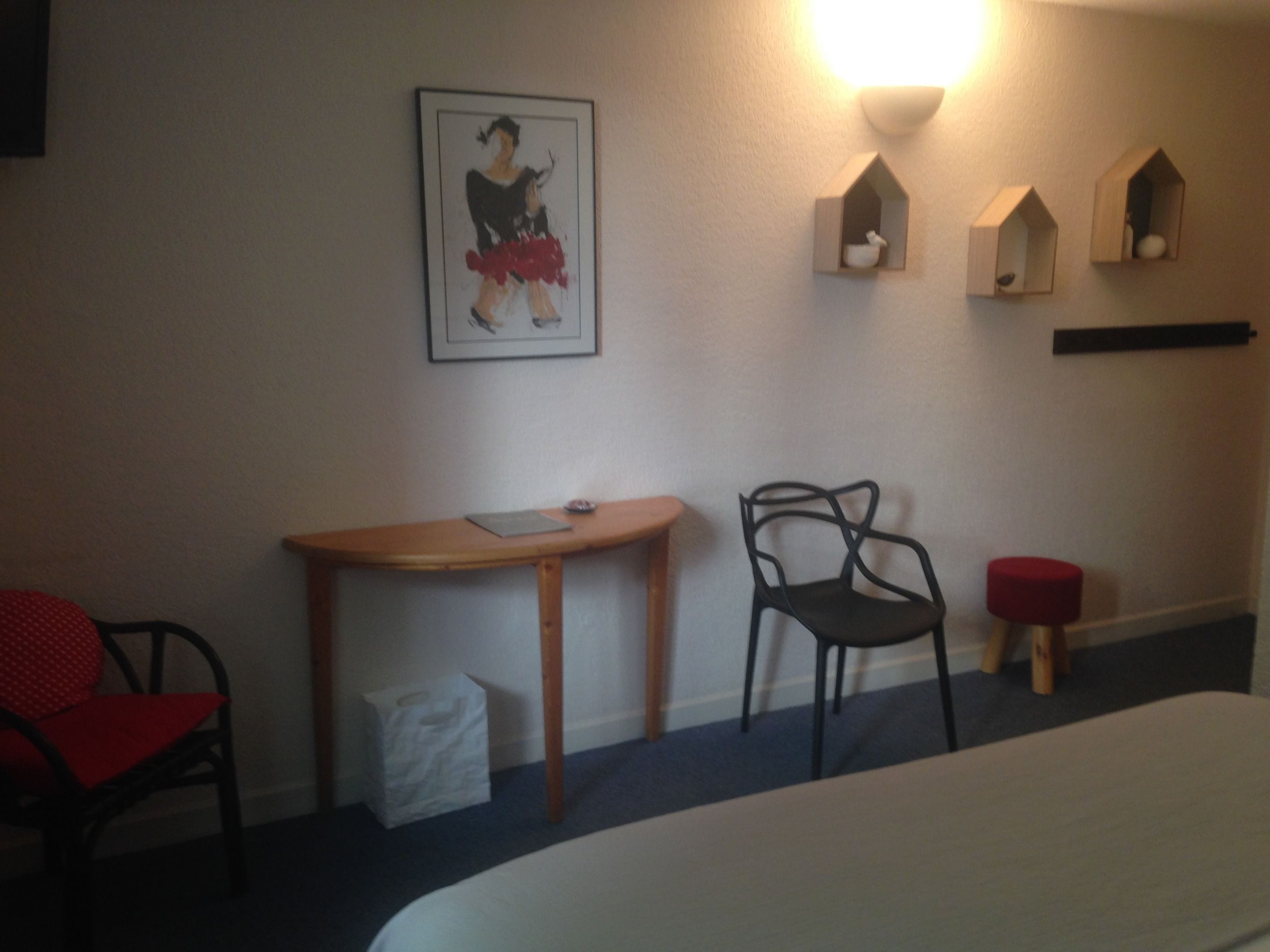 A hotel room with a bed, a desk, a chair, and a wall-mounted shelf with decorative items.