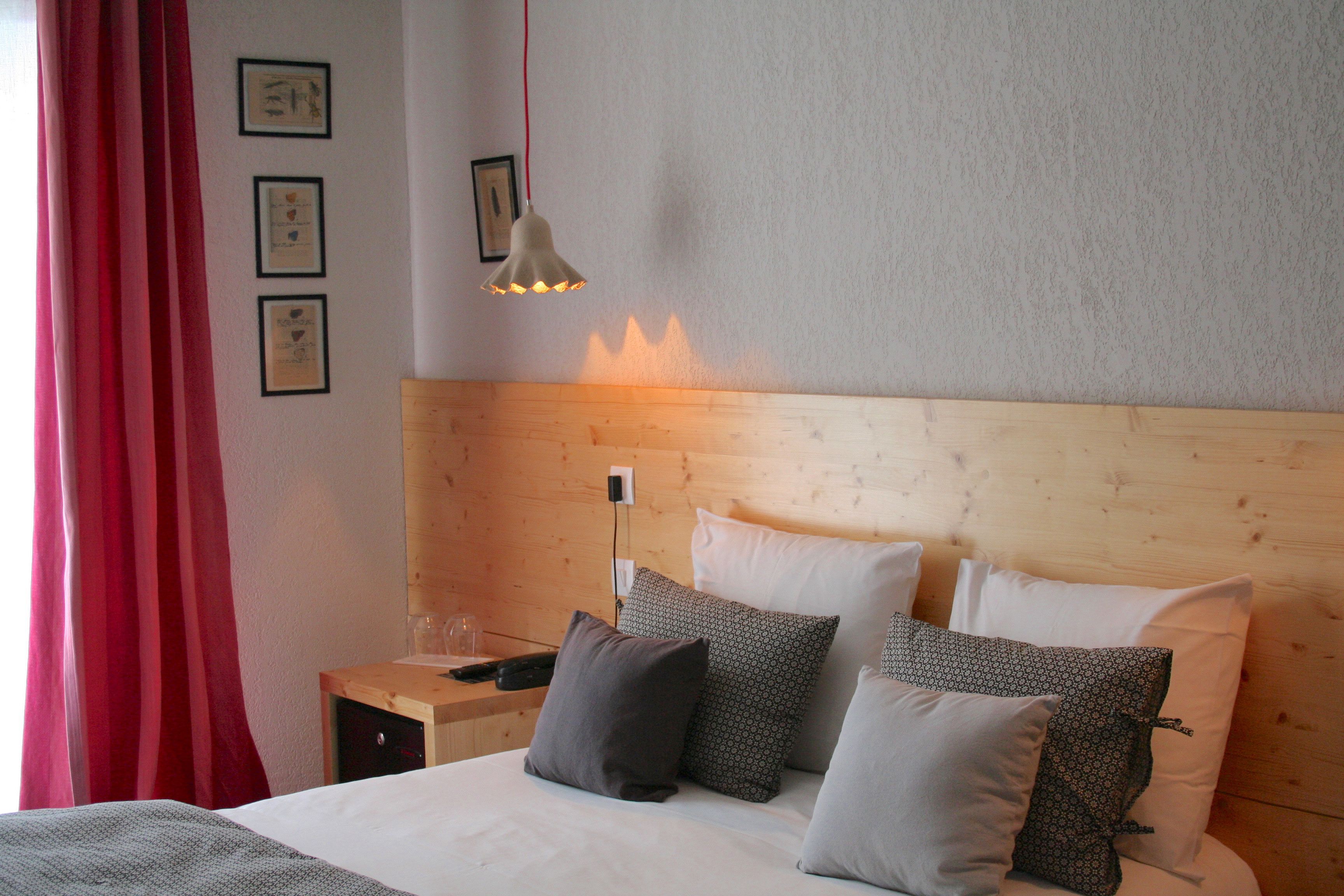 A neatly made bed with white and patterned pillows, a wooden headboard, a bedside table with a lamp, and a red curtain.