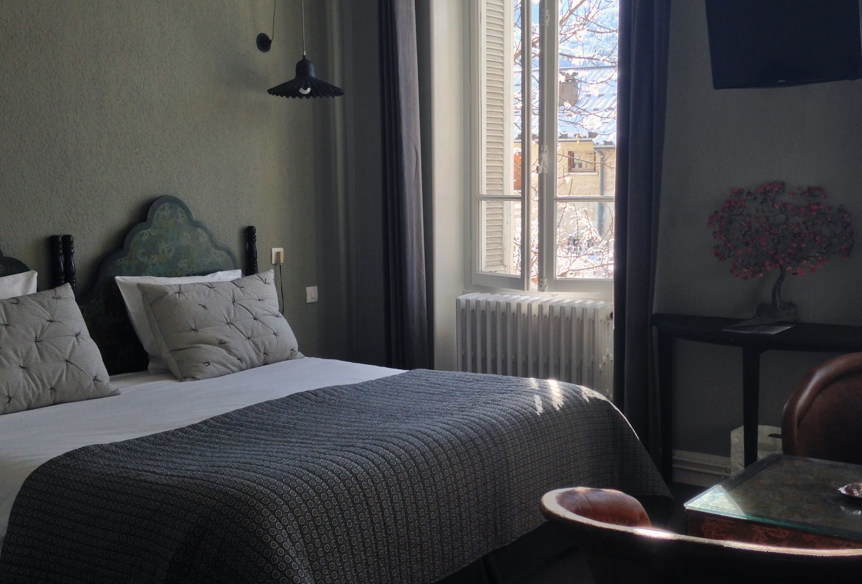A bedroom with a bed, a window with shutters, a radiator, a small table, and a decorative plant.