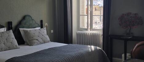 A bedroom with a bed, a window with shutters, a radiator, a small table, and a decorative plant.
