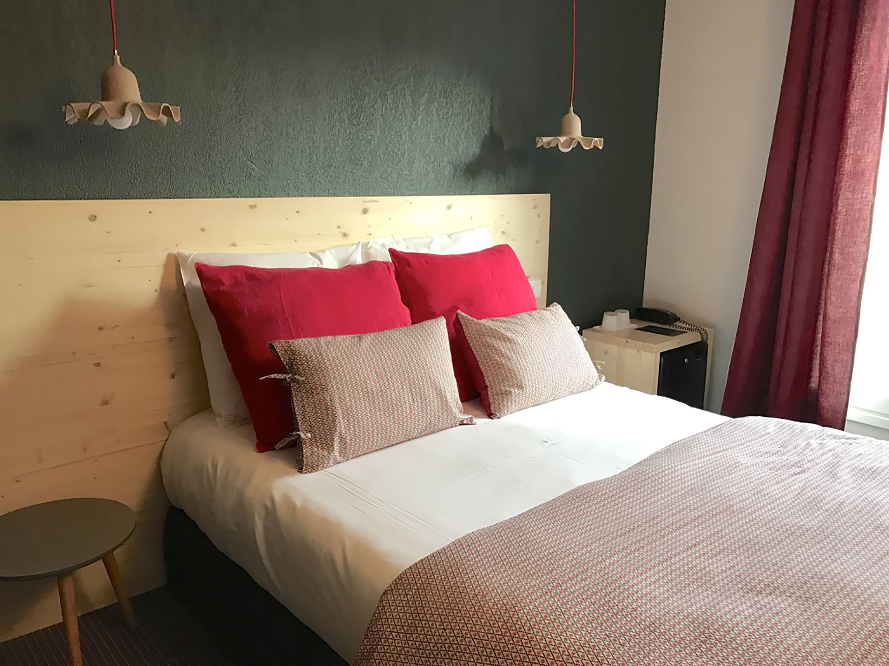 A hotel room with a wooden headboard, a bed with red and beige pillows, a bedside table, and a small round table.