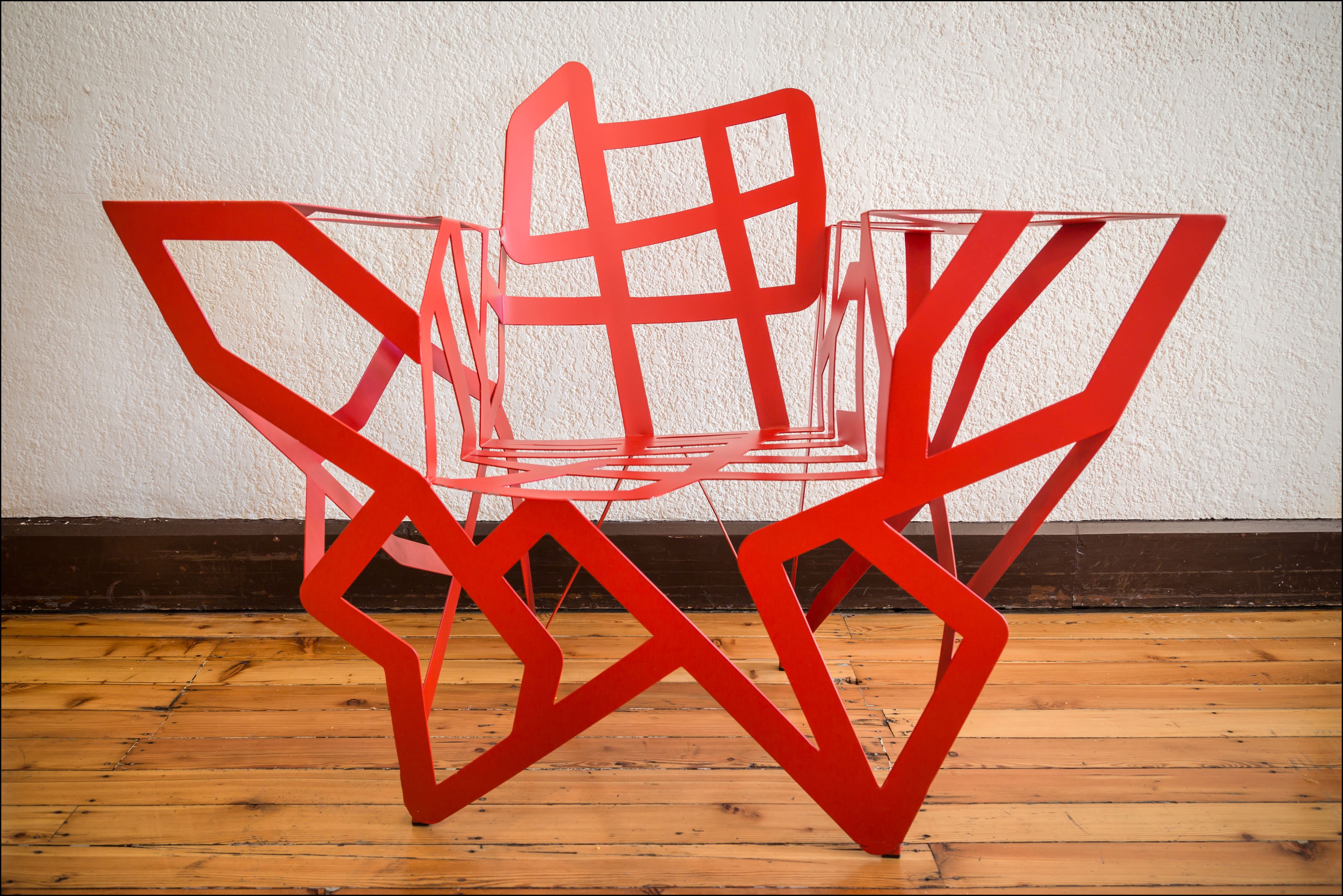 A red abstract chair with a geometric design, placed on a wooden floor against a white wall.