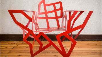 A red abstract chair with a geometric design, placed on a wooden floor against a white wall.