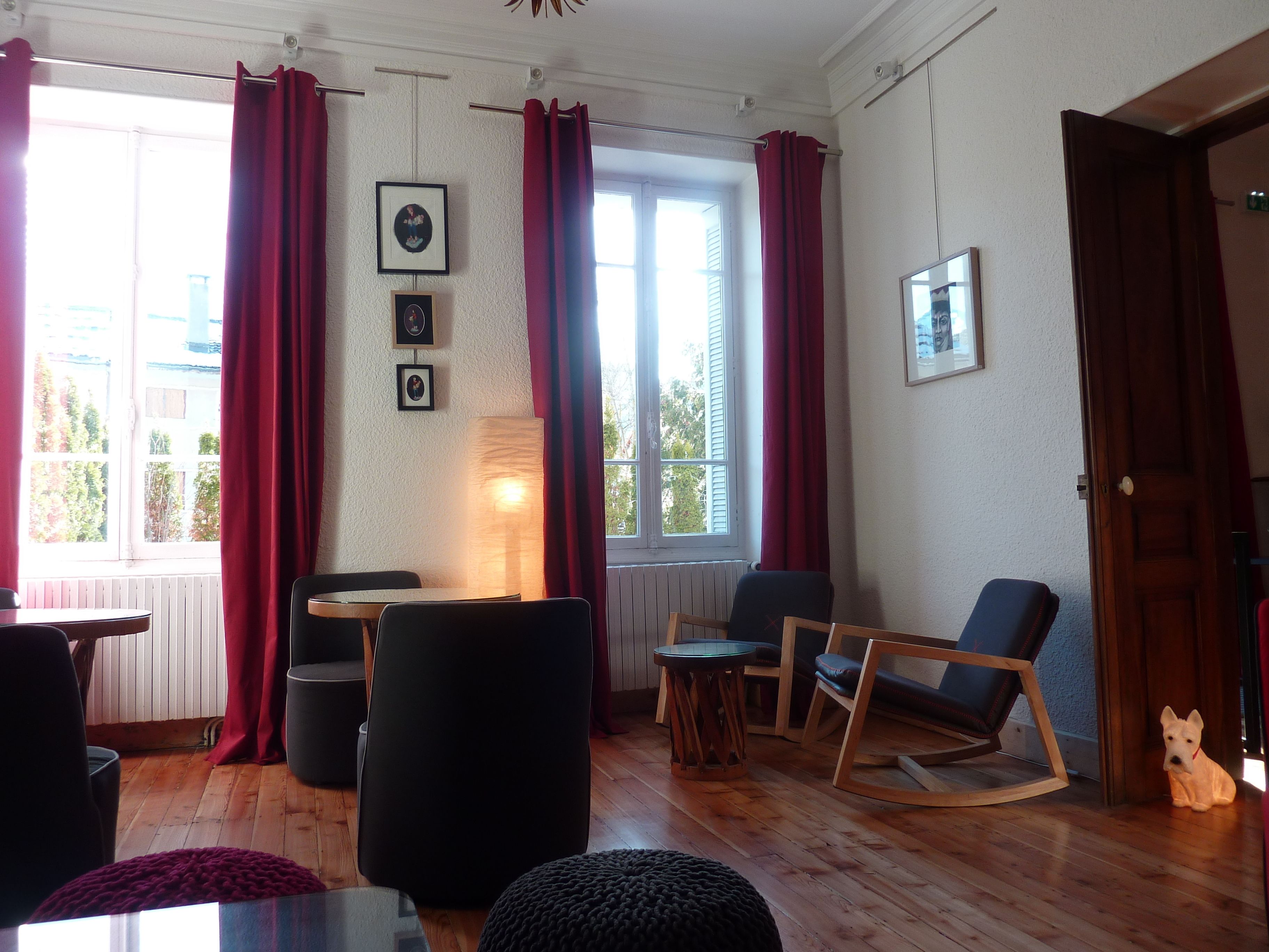 A cozy living room with a wooden floor, a dining table, a sofa, and a rocking chair. There are red curtains on the windows, and a lamp is on the table.