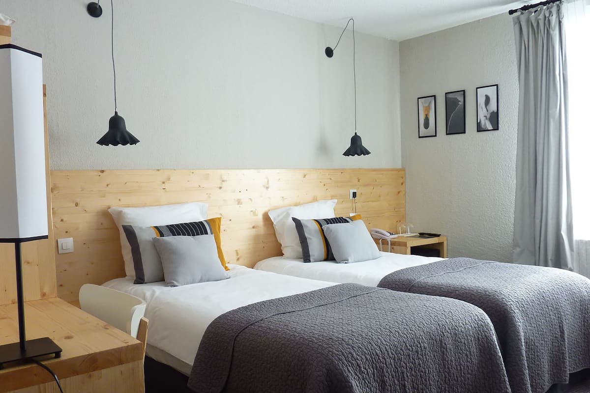 A hotel room with two beds, wooden headboards, a desk with a lamp, and a nightstand with a phone and glass.