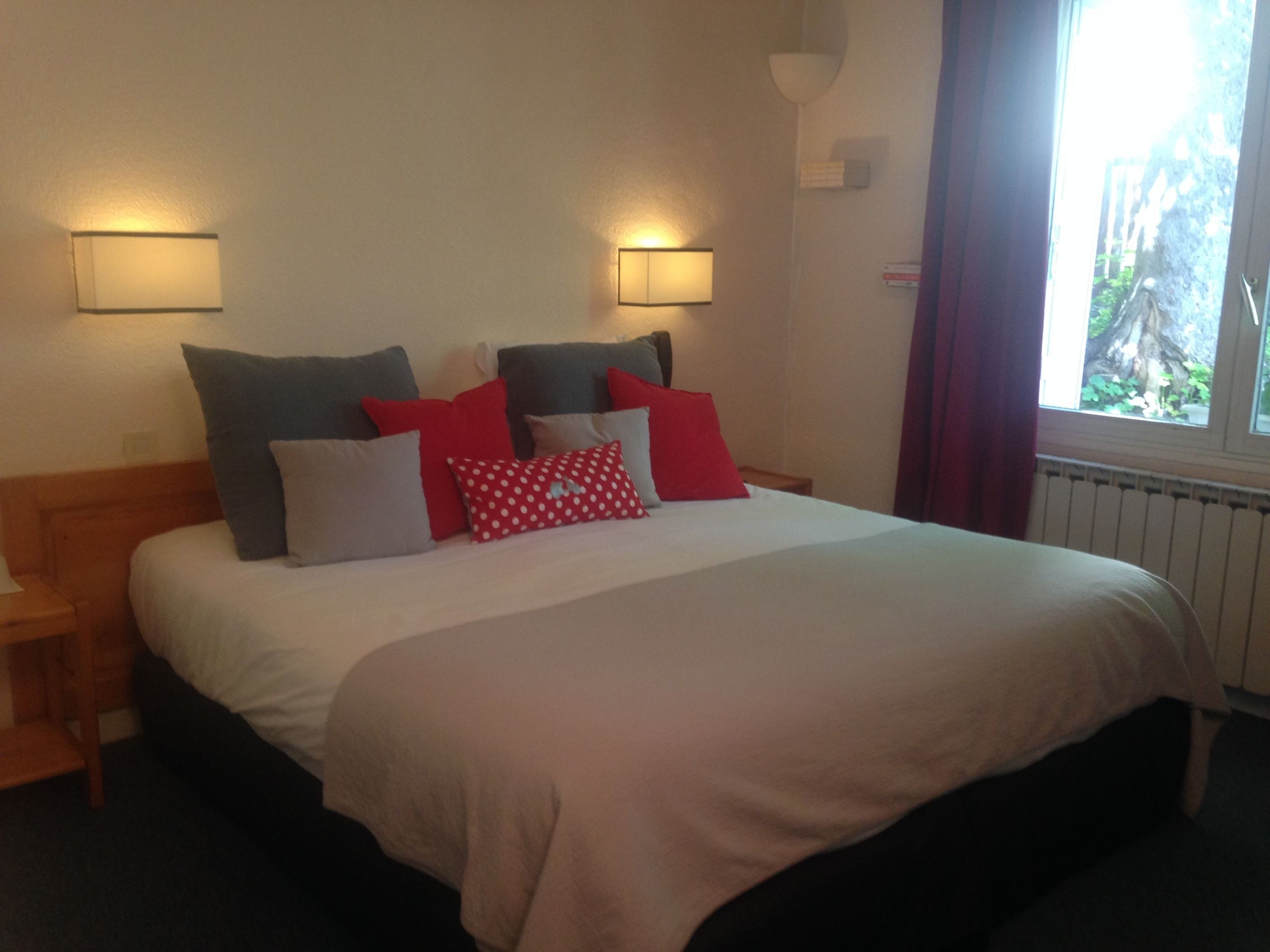 A hotel room with a large bed, two bedside lamps, a window with curtains, and a view of greenery outside.