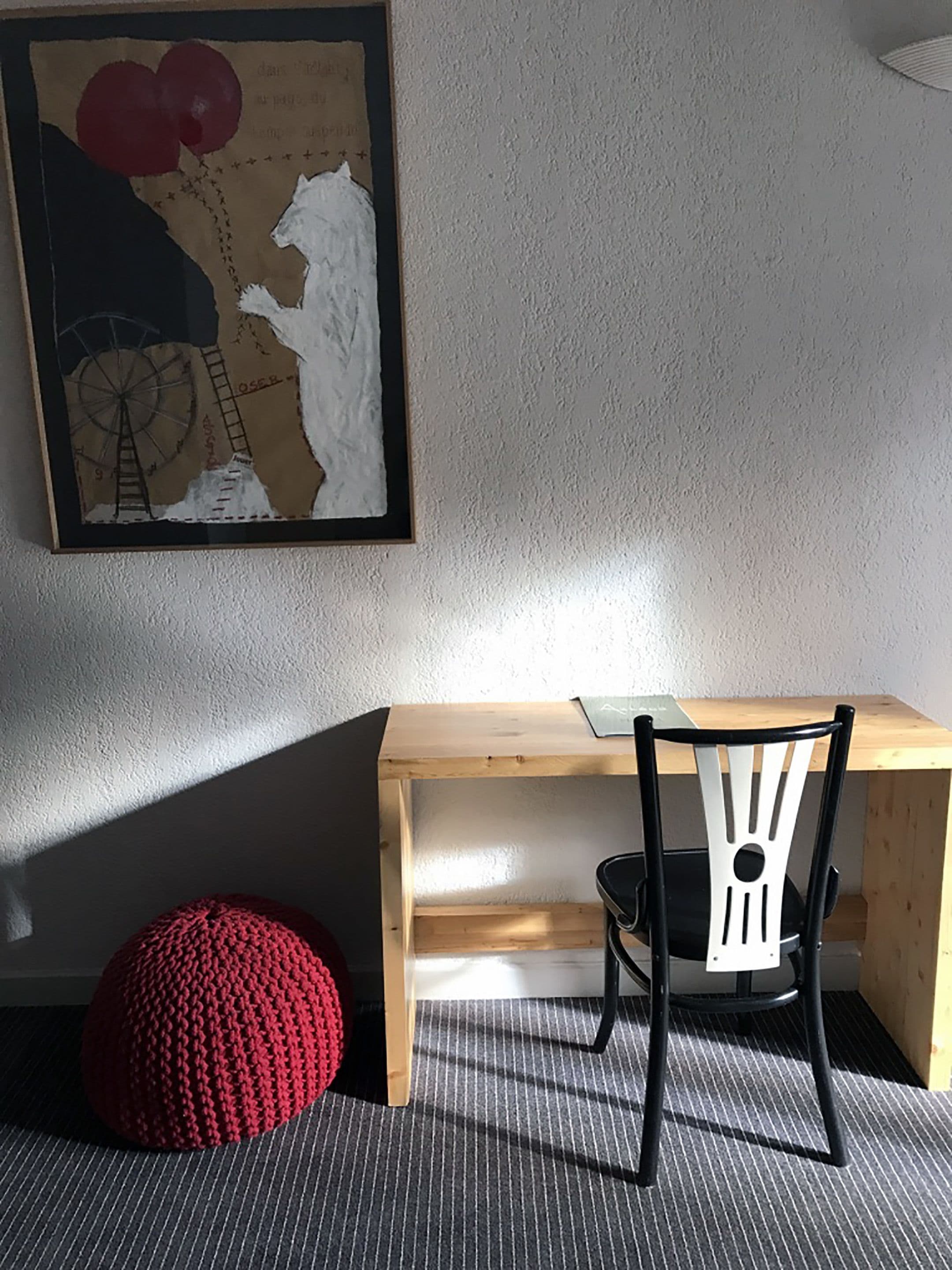 Ein Holztisch mit einem schwarzen Stuhl, einem roten Wollpouf und einem gerahmten abstrakten Gemälde an der Wand.