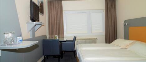 Habitación de hotel con cama, escritorio con silla, televisor montado en la pared y ventana con cortinas.