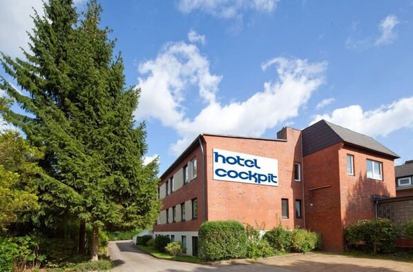 Un hôtel appelé « hôtel cockpit », avec une façade en briques, entouré de verdure et sous un ciel dégagé.