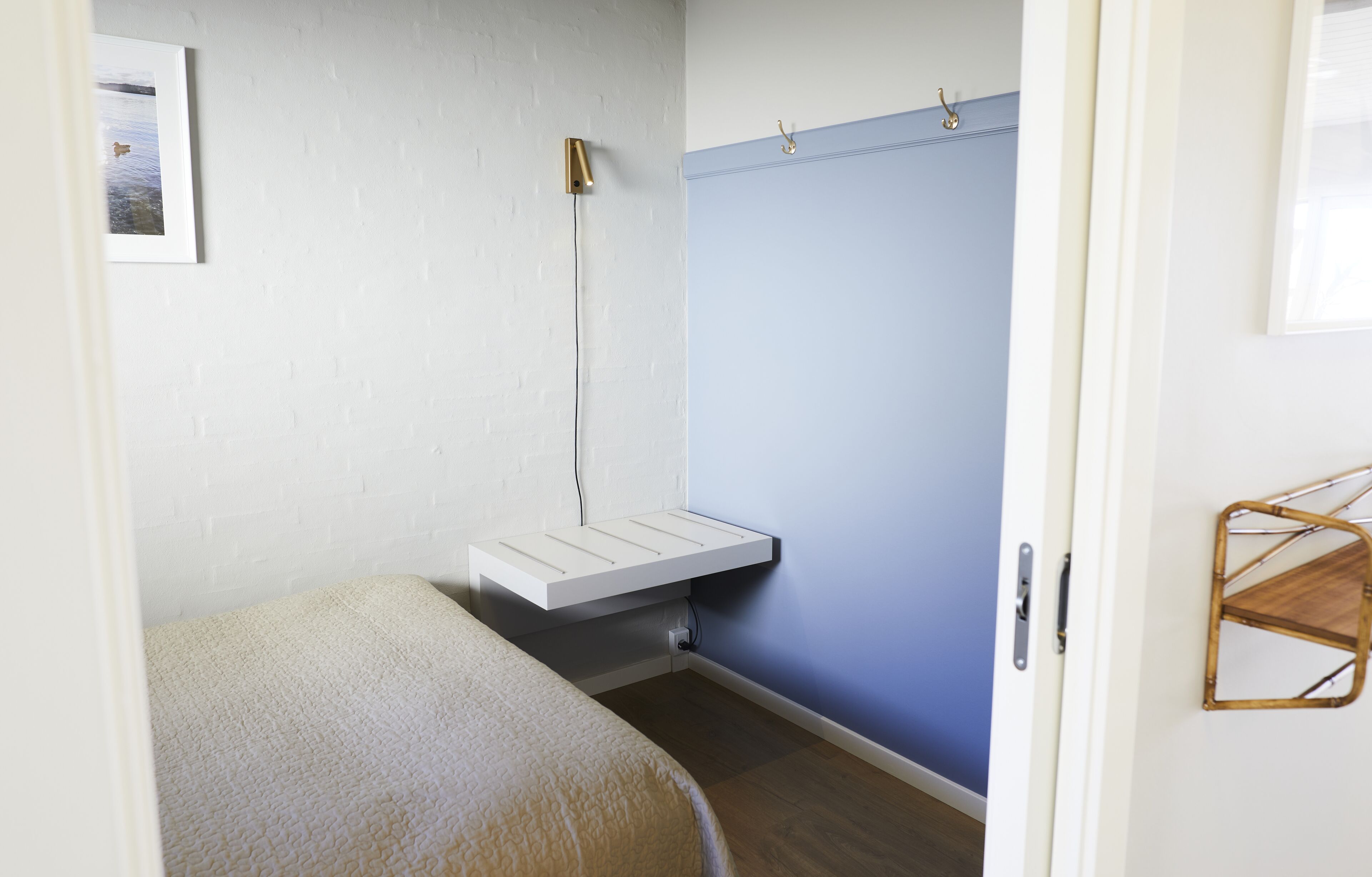 Una habitación pequeña con una cama, un estante blanco, una lámpara de pared y una puerta con manija.