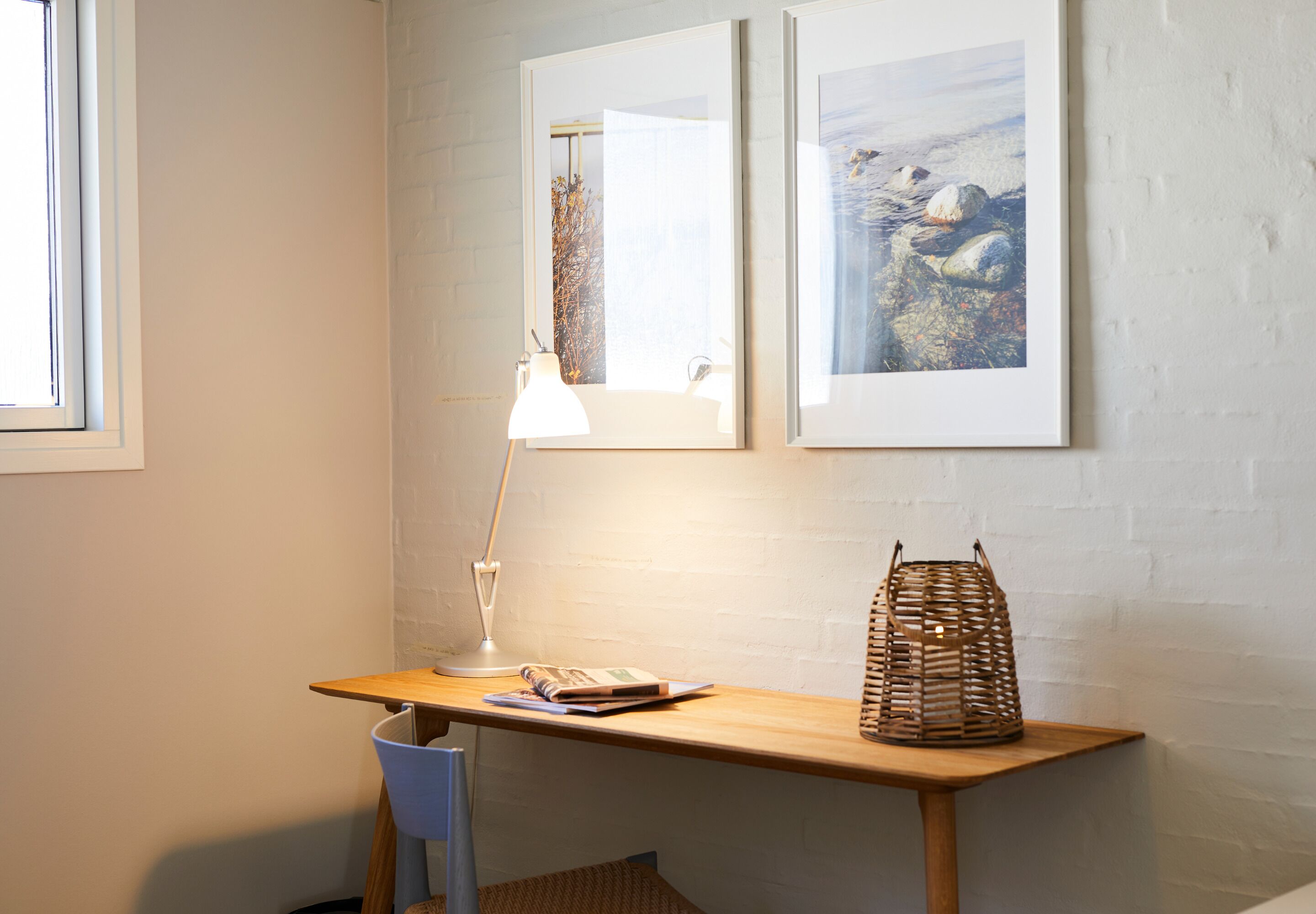 A room with a wooden desk, a chair, a lamp, a basket, and two framed pictures on the wall.