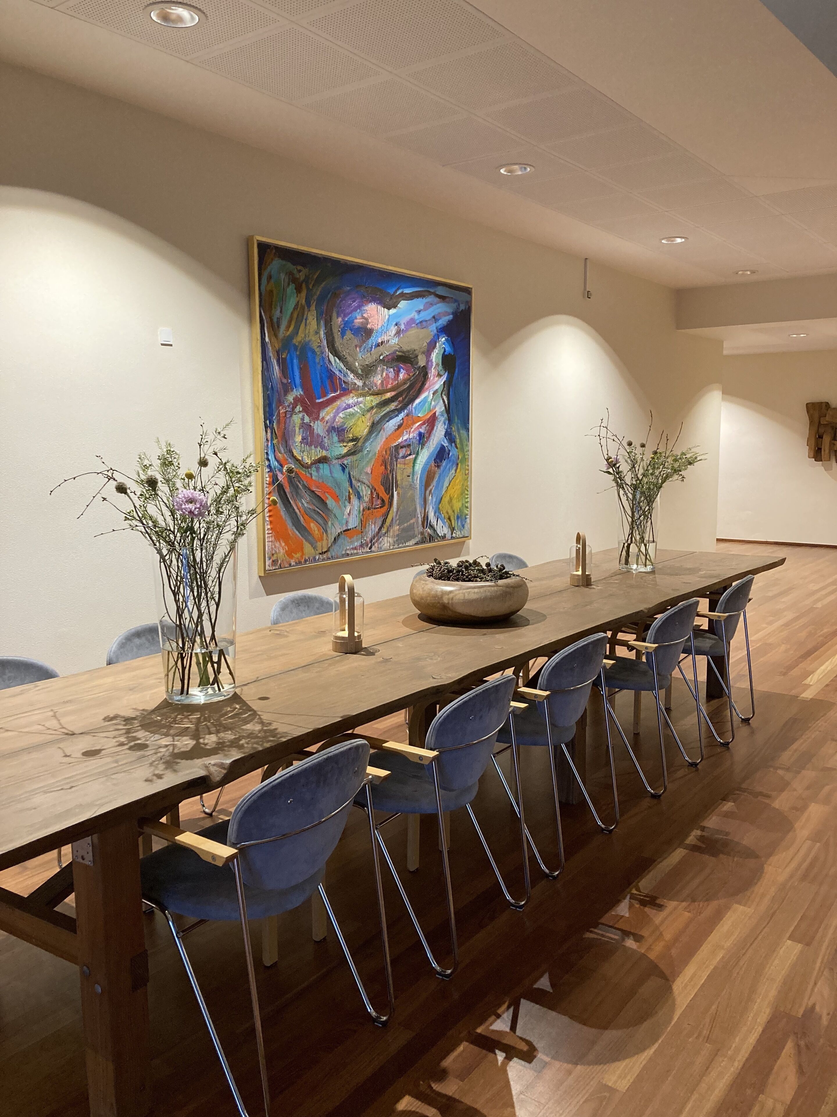Ein langer Holztisch für Besprechungen mit sechs blauen Stühlen, ein farbenfrohes abstraktes Gemälde an der Wand und je eine Vase mit Pflanzen auf jeder Seite.
