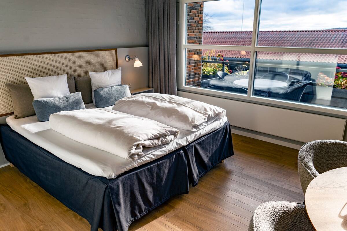 A neatly made bed with a headboard, a bedside table with a lamp, and a view of a red-tiled roof through a window.