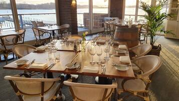 Restaurante con vista a un cuerpo de agua, mesas de madera dispuestas con vajilla y sillas de mimbre.