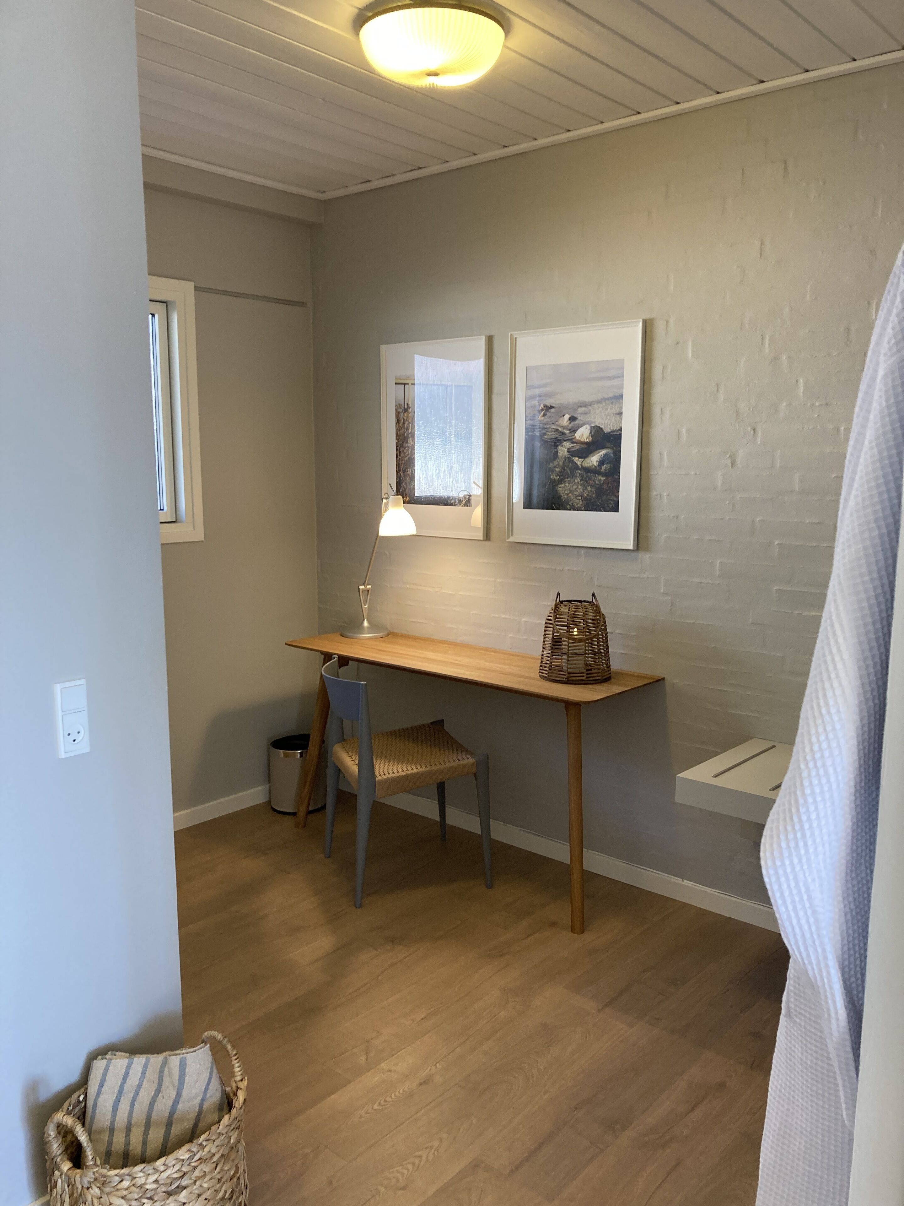 A small, well-lit room with a wooden desk, a chair, a lamp, and two framed pictures on the wall.