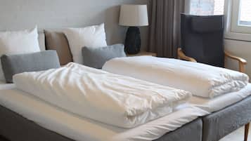 Uma cama de casal bem arrumada com lençóis brancos, uma mesinha de cabeceira com abajur e uma cadeira.