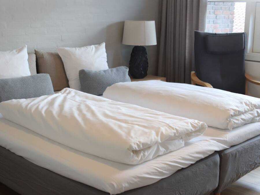 Uma cama de casal bem arrumada com lençóis brancos, uma mesinha de cabeceira com abajur e uma cadeira.