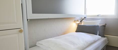 Pequeña habitación con cama individual, ropa de cama blanca, cabecera blanca y ventana con luz natural.