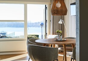 A dining area with a round table, grey upholstered chairs, a hanging light fixture, and a view of a body of water through large windows.